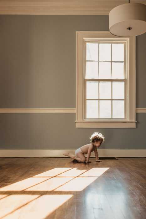 toddler in diaper crawling across light hardwood floor in an empty room under window with light pouring in family photo session